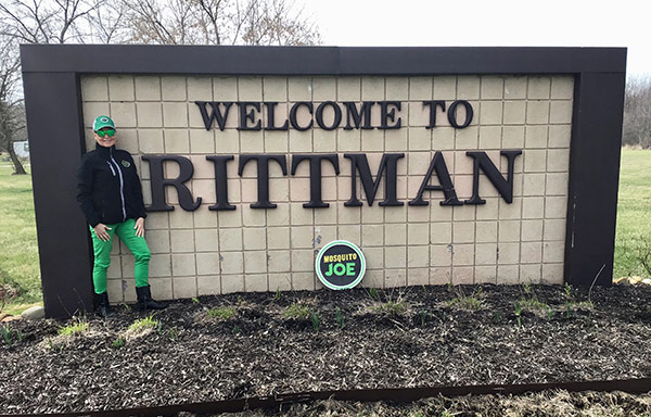 Lady in a black and green Mosquito Joe outfit poses for picture next to Welcome to Rittman is sign in Rittman, OH